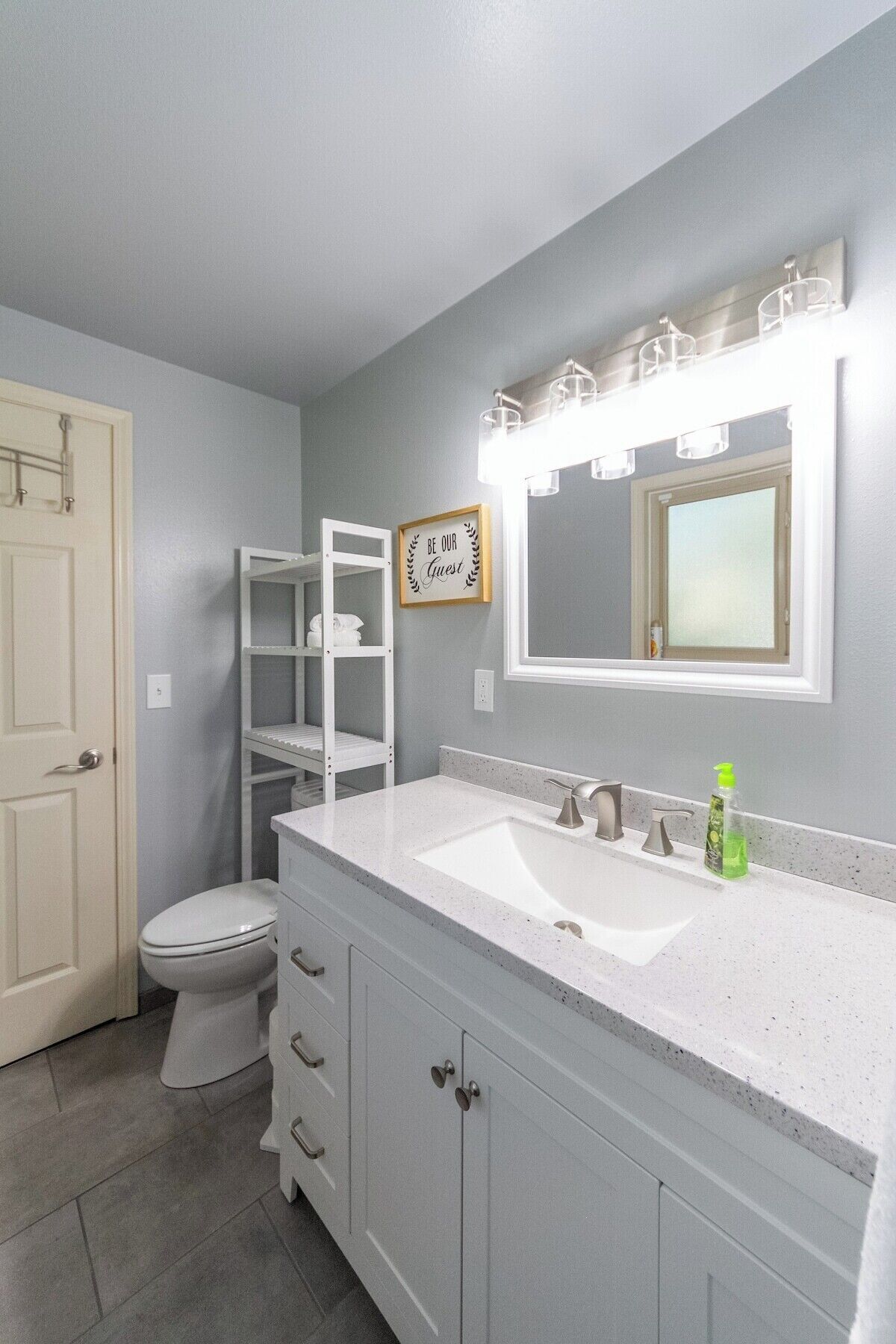 A bathroom with a white sink, a toilet, and a vanity with drawers. There is a mirror with lights above it and a sign that says "Be Our Guest".