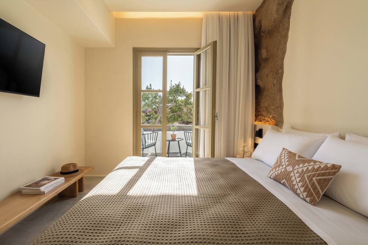 A bedroom with a large bed, a flat-screen TV, a wooden bench, and a view of the outdoors through a sliding door.
