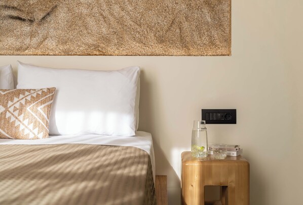 Una cama bien hecha con almohadillas estampadas, una mesita de noche de madera con un vaso de agua y libros, y un tapiz con textura en la pared de arriba.