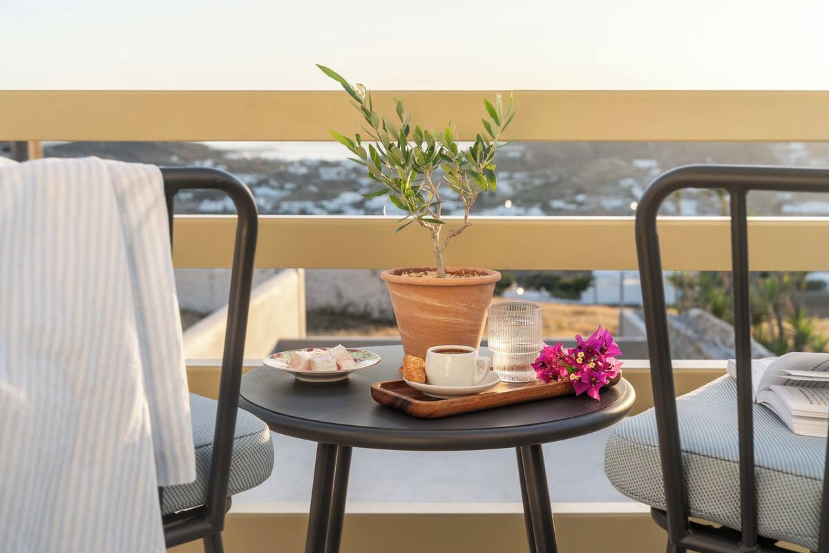 A balcony with a potted plant, a small table with a cup of coffee, a plate of food, and a glass of water.