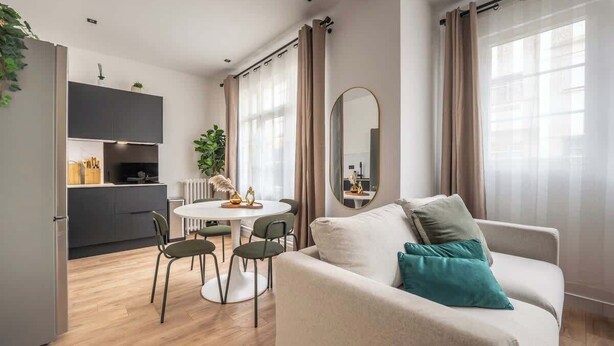 Ein modernes Wohnzimmer mit Sofa, Esstisch mit Stühlen, einer Küchenzeile und großen Fenstern mit Vorhängen.