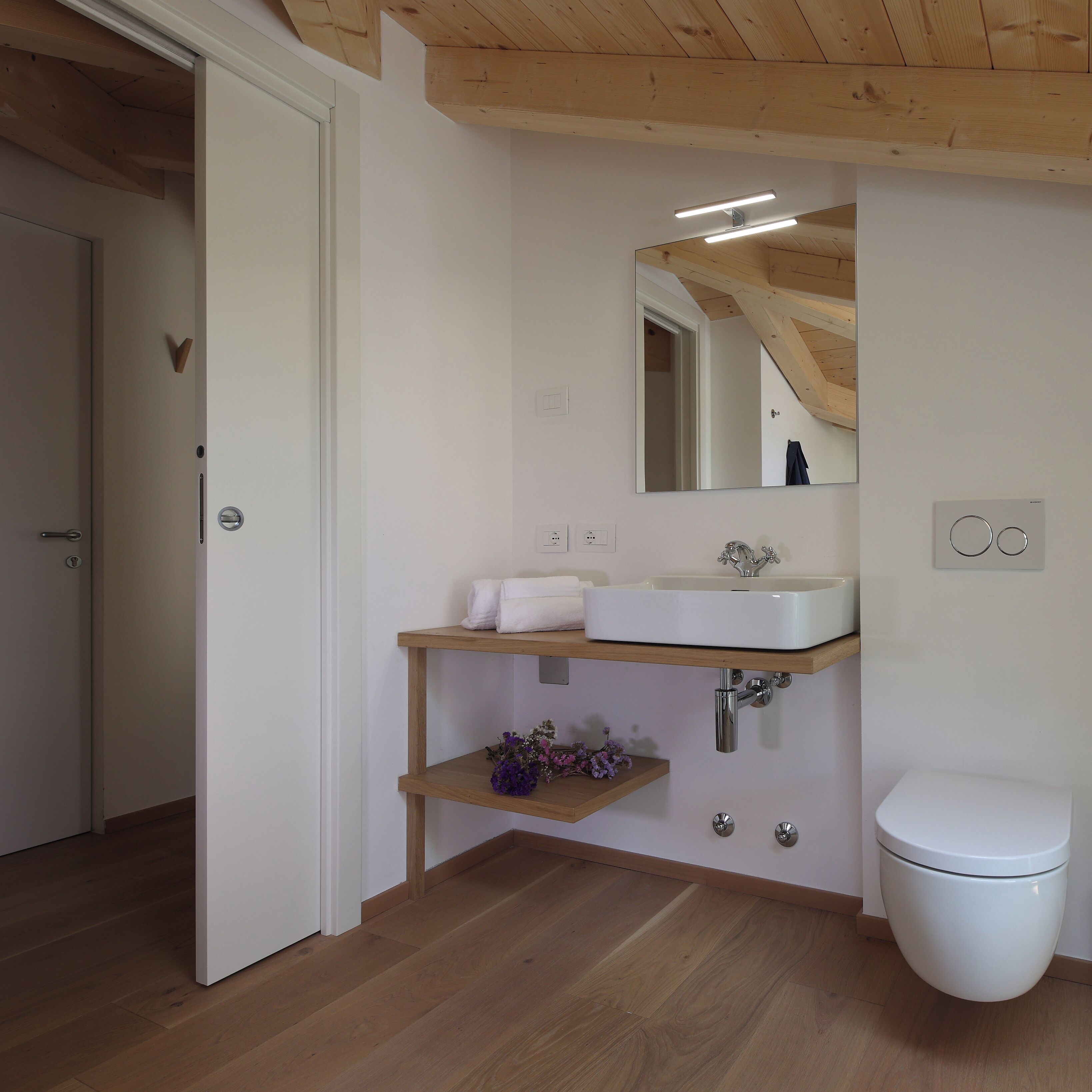 A modern bathroom with a wooden floor, white walls, a white sink, a mirror, and a toilet.