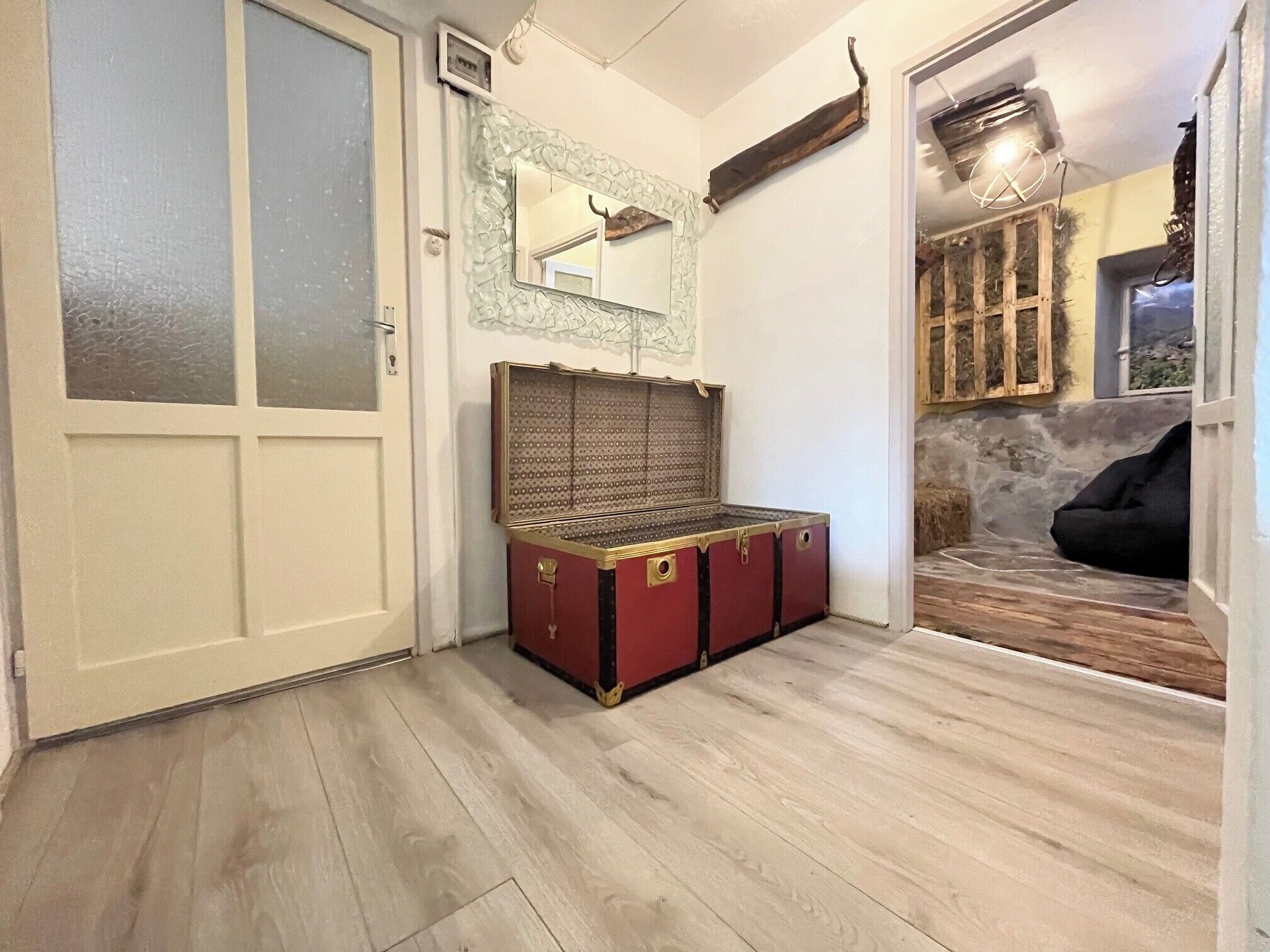 A room with a wooden floor, a red and black trunk, a white door, and a mirror with a stone frame.
