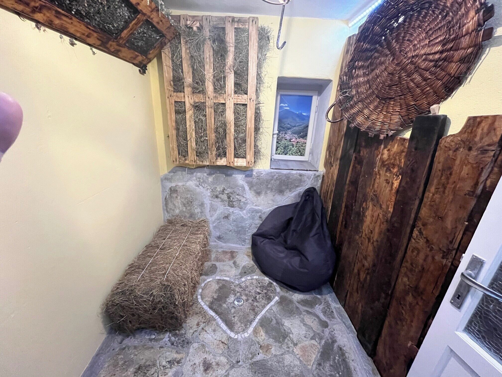 Un banco de piedra con diseño en forma de corazón, un sillón de cuero negro y un fardo de heno en una habitación rústica con una puerta de madera y una ventana con vistas a la montaña.