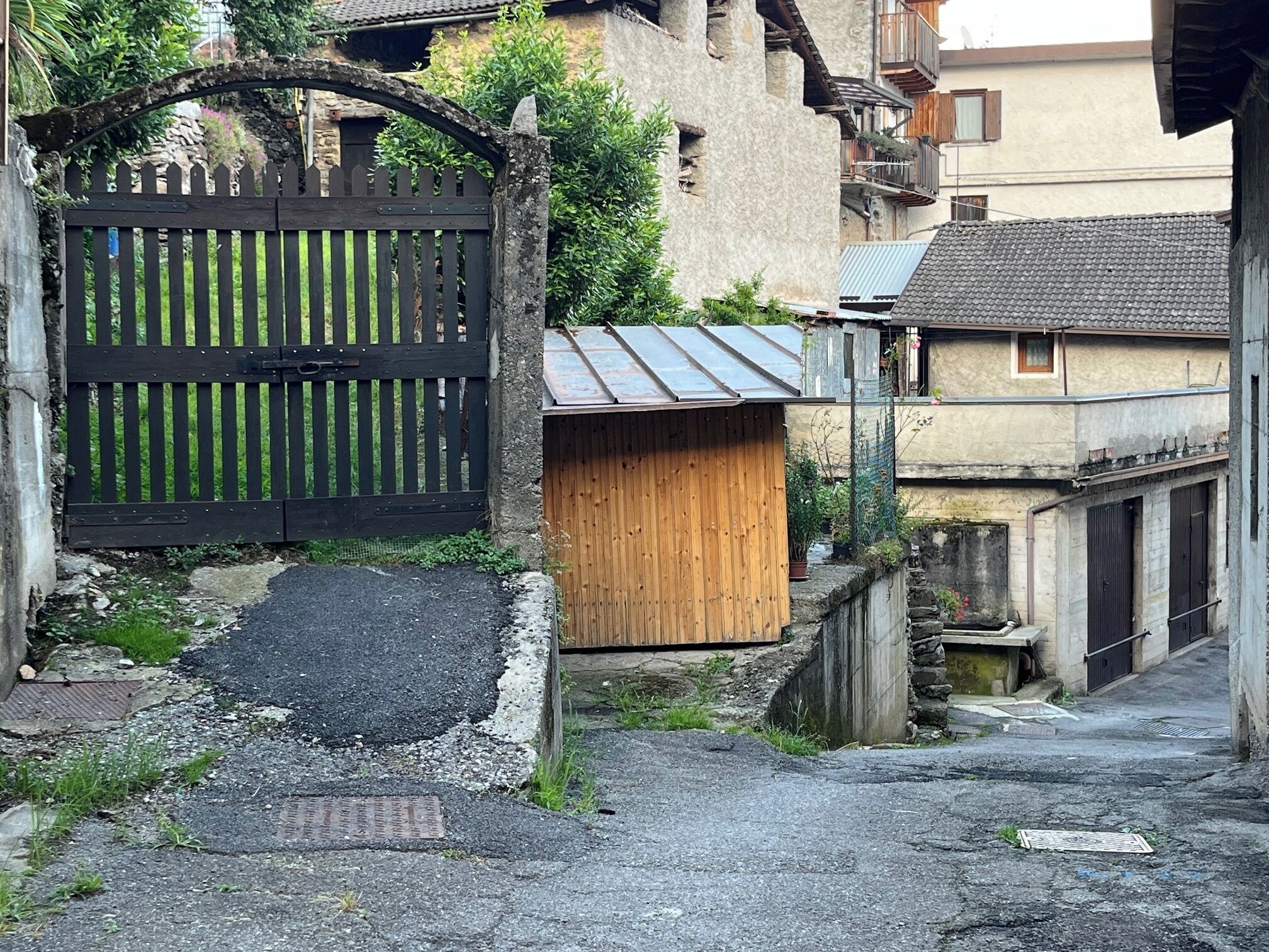 Un stretto vicolo con un cancello in legno, una piccola costruzione in legno e un edificio con tetto in tegole.