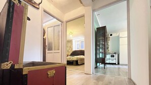 A room with a large red suitcase, a bed with a patterned bedspread, a dresser with a glass display case, and a kitchen area with white cabinets. - Sorriso Berzo Apartment With Stunning Views (Berzo Demo)