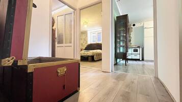 Una stanza con una valigia rossa grande, un letto con copripiumino fantasia, una cassettiera con vetrina e un angolo cottura con mobili bianchi.