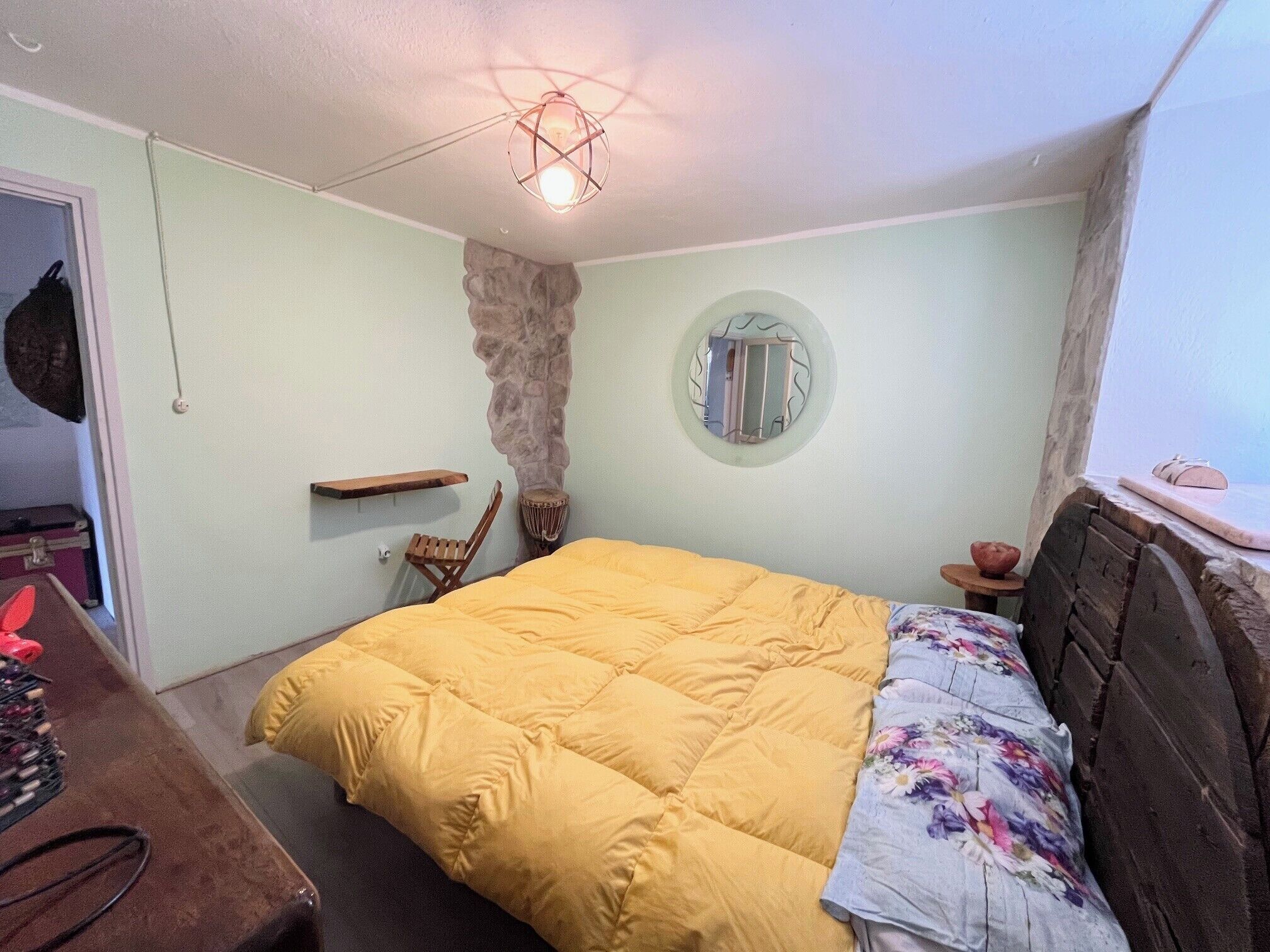 A bedroom with a bed, a wooden headboard, a stone accent wall, a round mirror, and a hanging light fixture.
