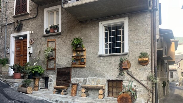 A rustic stone building with wooden benches, potted plants, and a wooden door. - Sorriso Berzo Apartment With Stunning Views (Berzo Demo)