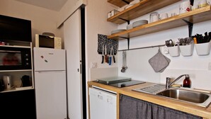 Uma cozinha compacta com geladeira branca, micro-ondas, lava-louças e pia. Há prateleiras de madeira acima do balcão com louças e utensílios.