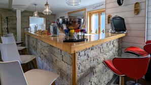 A kitchen with a wooden bar, stone walls, and red bar stools. There are hanging lights, a large bowl, and a television on the bar. - Spacious family chalet with spa, sauna and games room (Ban-sur-Meurthe-Clefcy)