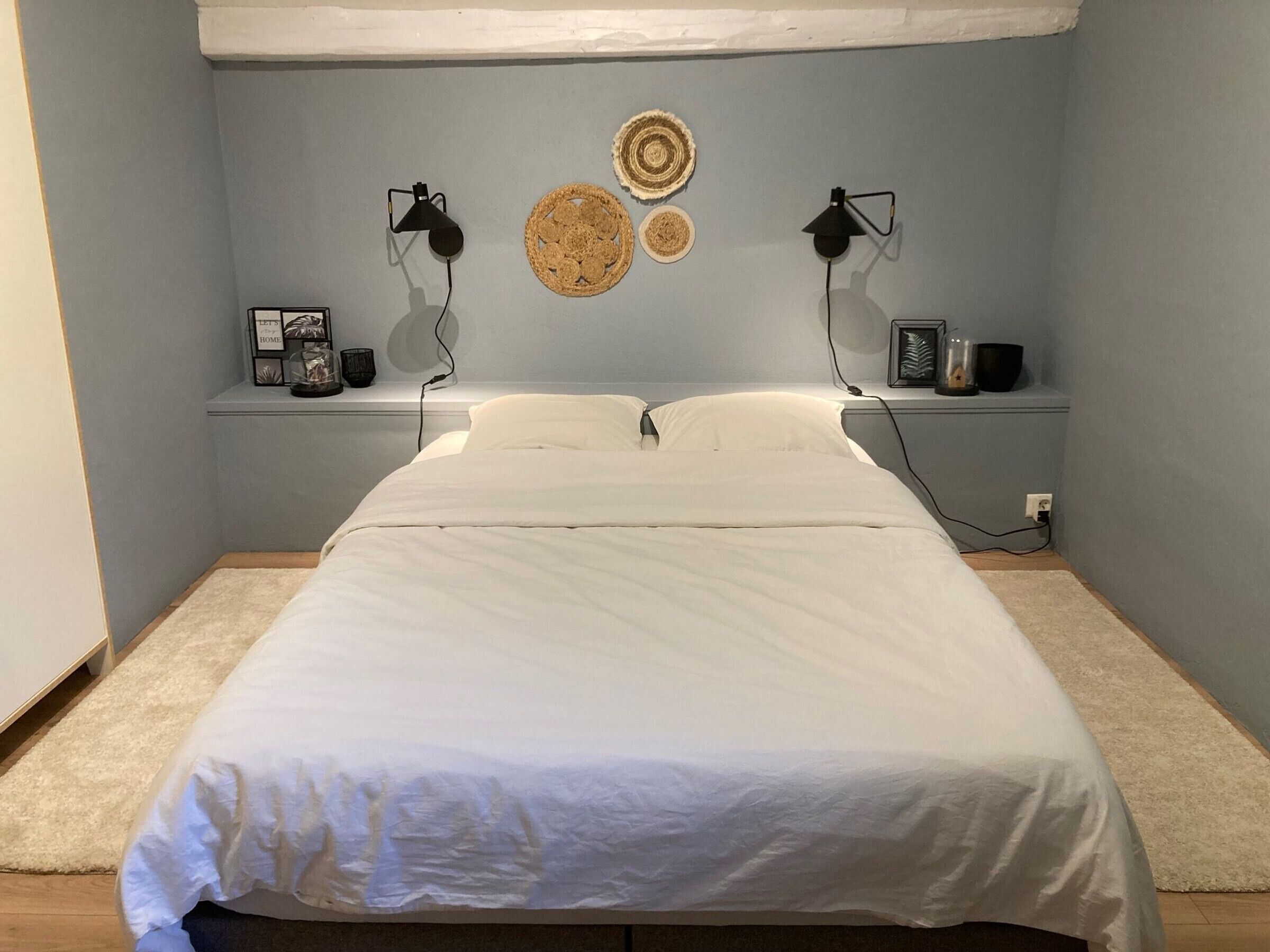 A bedroom with a bed, two bedside lamps, a shelf with decorative items, and a wall-mounted light fixture.