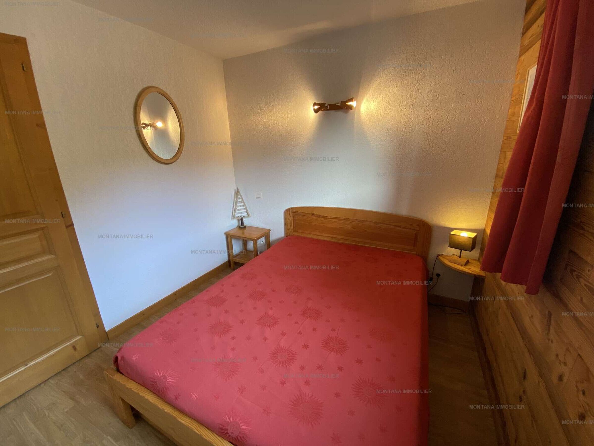 Un dormitorio con una cama de madera, una mesita de noche con lámpara, un espejo en la pared y una puerta.