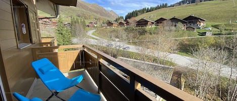 A balcony with two blue chairs, a wooden railing, and a view of a valley with houses and mountains.