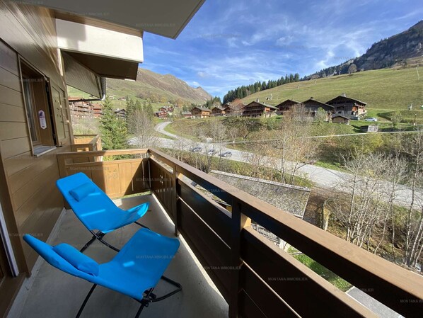 A balcony with two blue chairs, a wooden railing, and a view of a valley with houses and mountains.