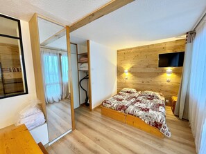 A bedroom with a wooden headboard, a bed with patterned bedding, a TV mounted on the wall, and a closet with a mirror.