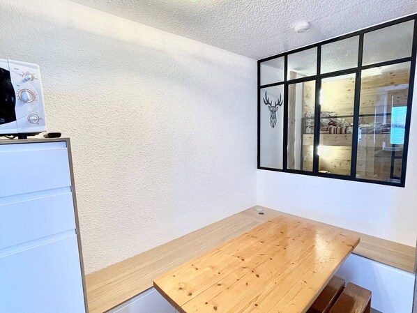 A small kitchenette with a microwave, a wooden table, and a window with a decorative headboard.