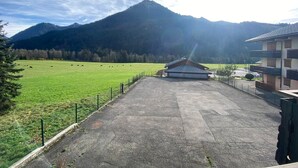 Un espace de stationnement dallé avec un bâtiment au centre, entouré d’un champ verdoyant clôturé où paissent des vaches, et des montagnes en arrière-plan.