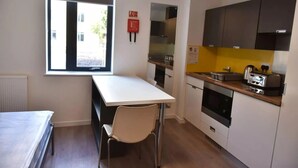 Et lille køkken med en hvid bordplade, en brødrister, og en komfur. Der er et vindue der giver naturligt lys og en stol foran et lille bord.