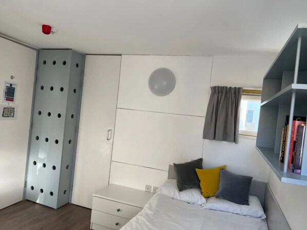 A bedroom with a bed, a wardrobe, a shelf with books, and a window with curtains. - Pittville Student Village (Cheltenham)