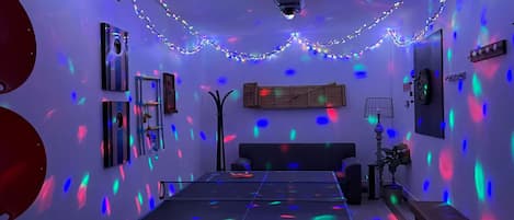 A room with a ping pong table, a wooden headboard, a wall-mounted clock, and string lights.