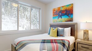 A bedroom with a large window, a bed with multiple pillows, a colorful painting above the bed, and a nightstand with a lamp.