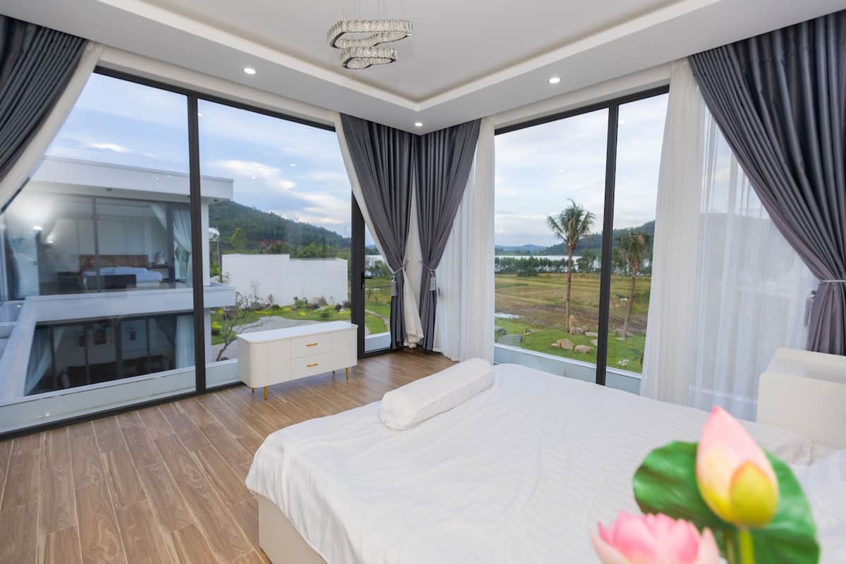 A modern bedroom with a large bed, a nightstand, and a view of a lush landscape through large windows.