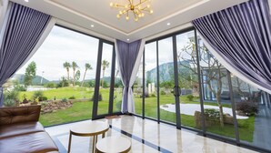 A spacious room with a large window, a modern chandelier, and a view of a landscaped garden. - TKH Resort (Hoa Son)