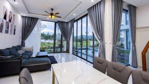 A modern living room with a sofa, a dining table, and large windows offering a view of the outdoors. - TKH Resort (Hoa Son)