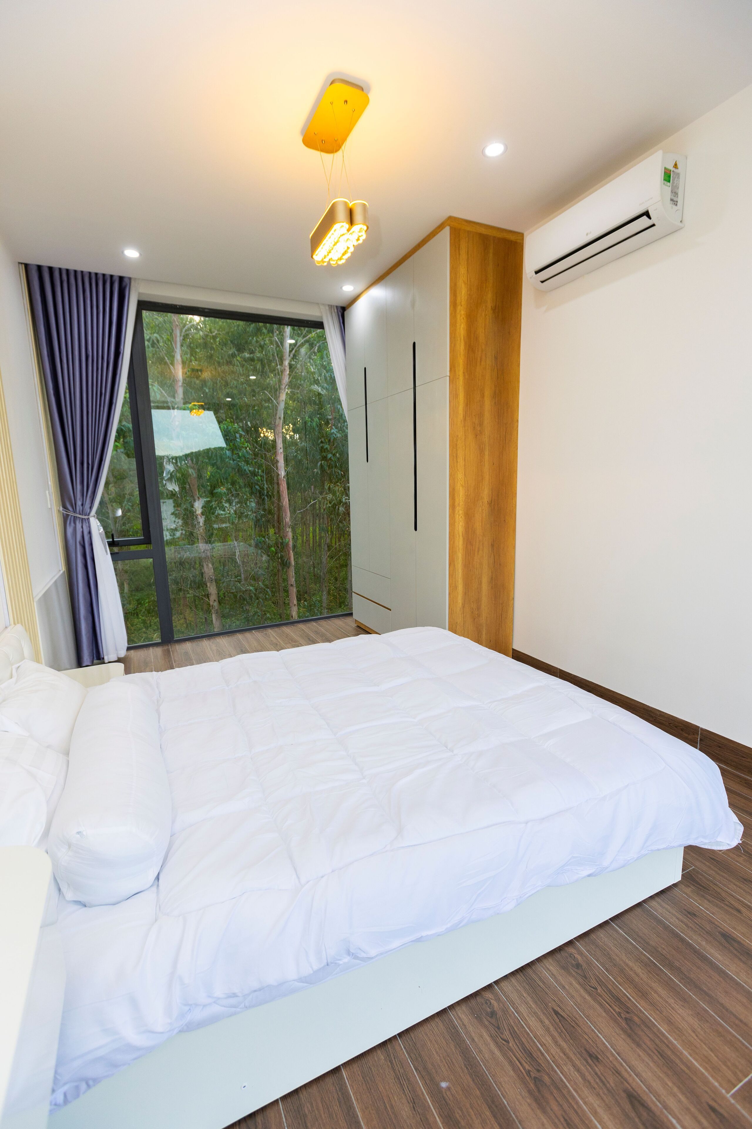 A modern bedroom with a large bed, a wooden accent wall, a window with curtains, and an air conditioning unit.
