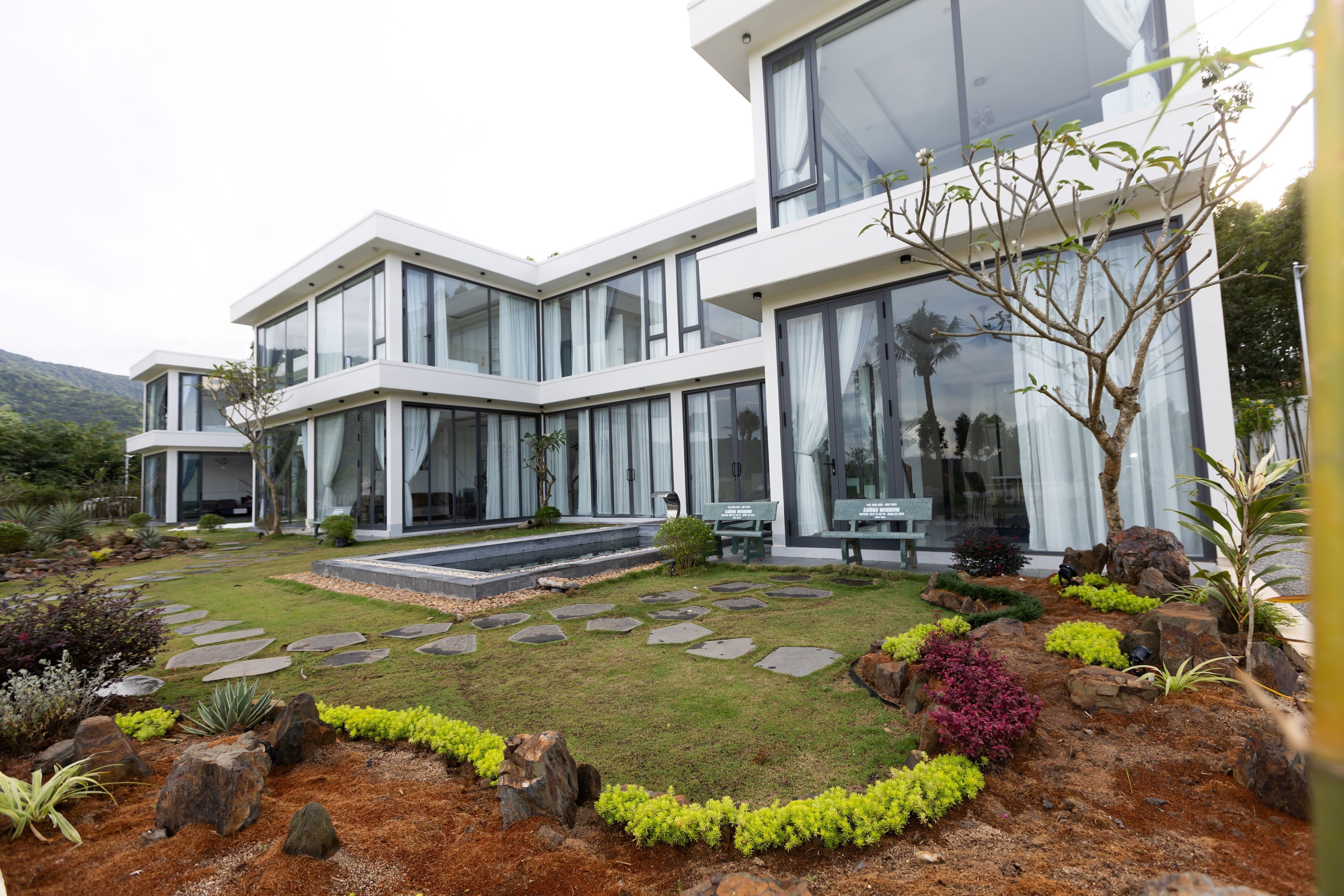 A modern two-story house with large windows, a stone pathway, and a landscaped garden with rocks and shrubs.