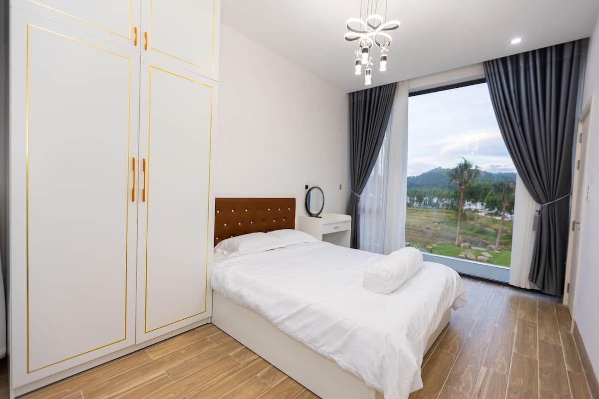 A bedroom with a bed, a wardrobe, a bedside table, a mirror, and a view of a landscape through a window.