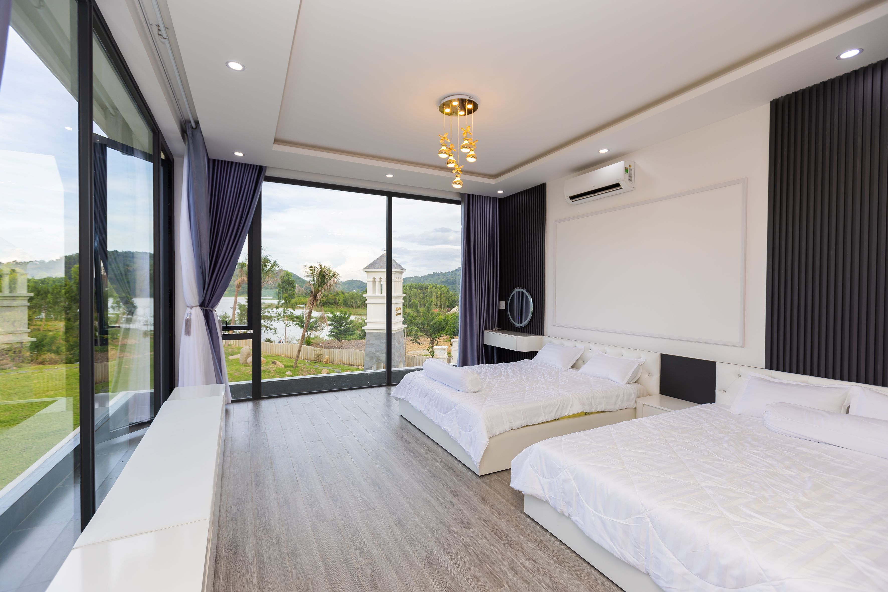 Quarto de hotel moderno com duas camas, uma janela grande com vista para área verde e um prédio, e um lustre no teto.