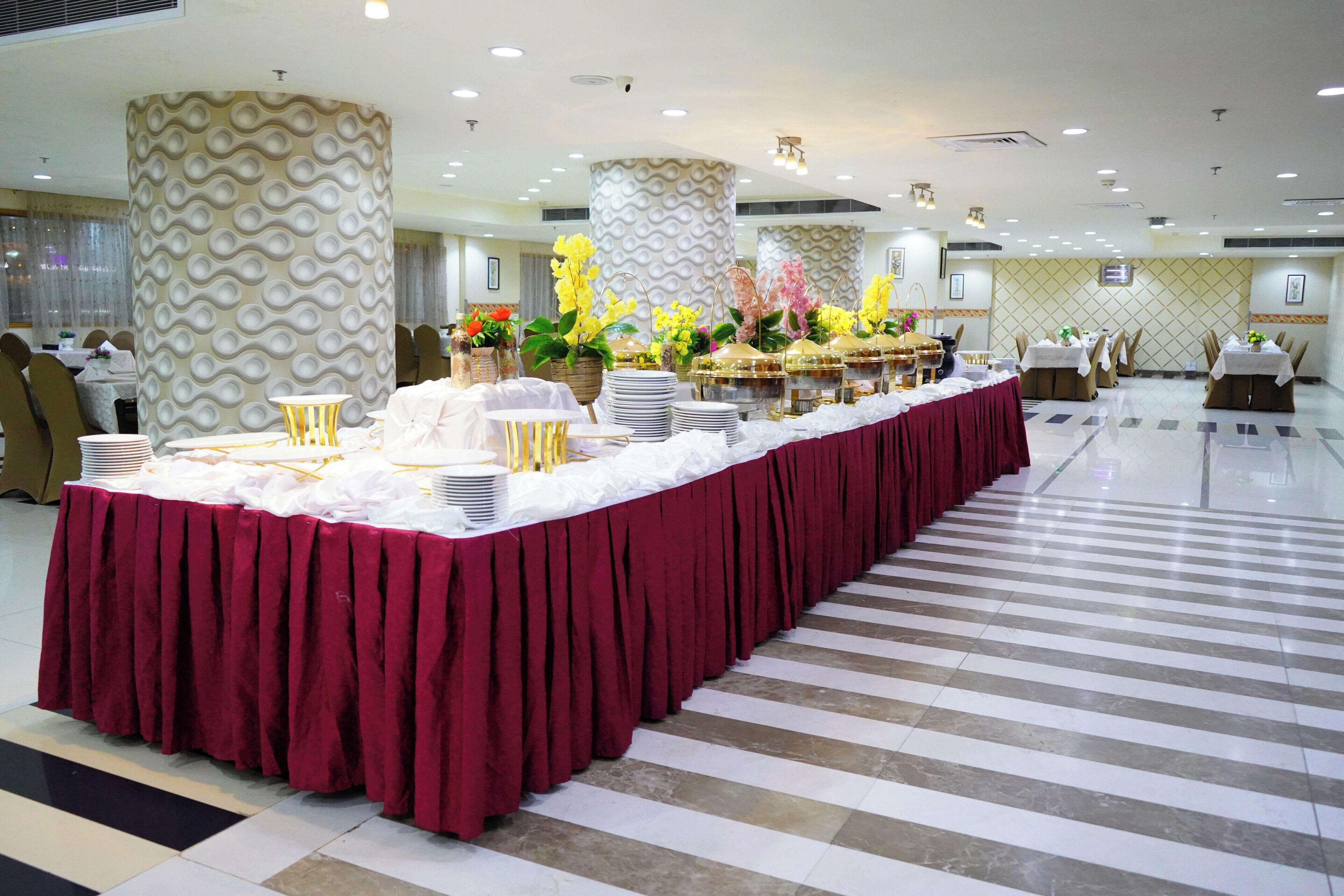 A banquet hall with a long table covered in a red tablecloth, surrounded by chairs and decorated with floral arrangements.