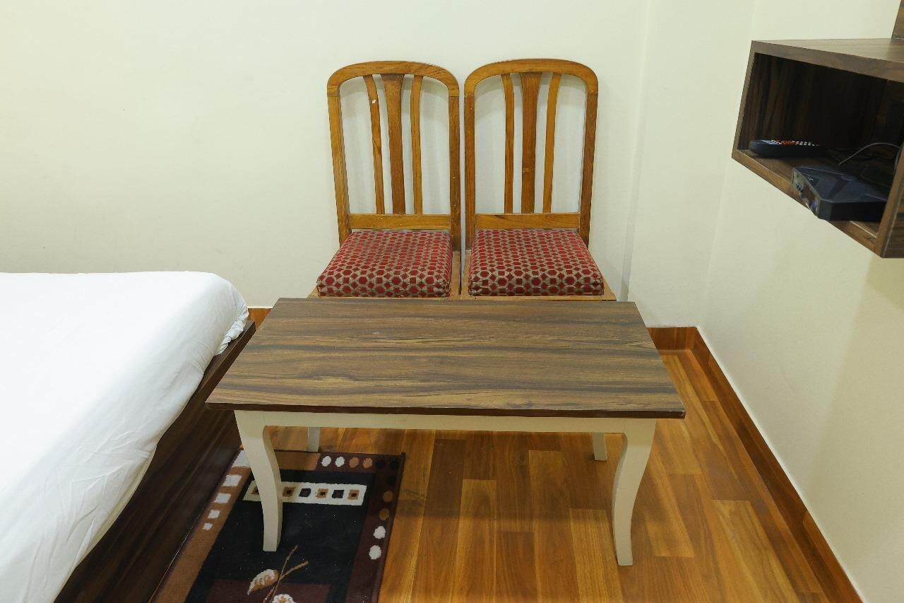 Een kleine kamer met een bed, een houten tafel, twee stoelen met gestreepte kussens en een plank met een apparaat.