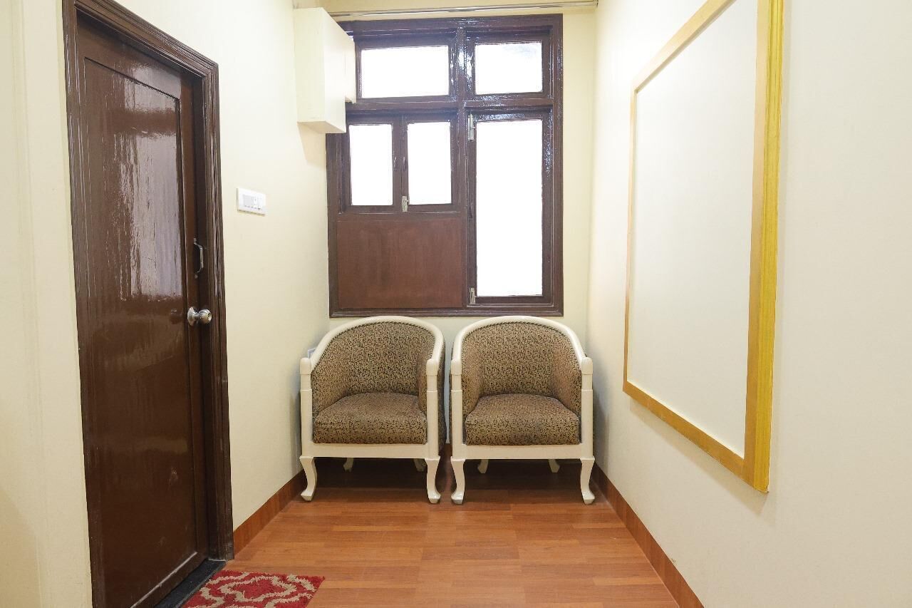 A narrow hallway with a wooden door, two patterned chairs, a window with multiple panes, and a framed picture on the wall.