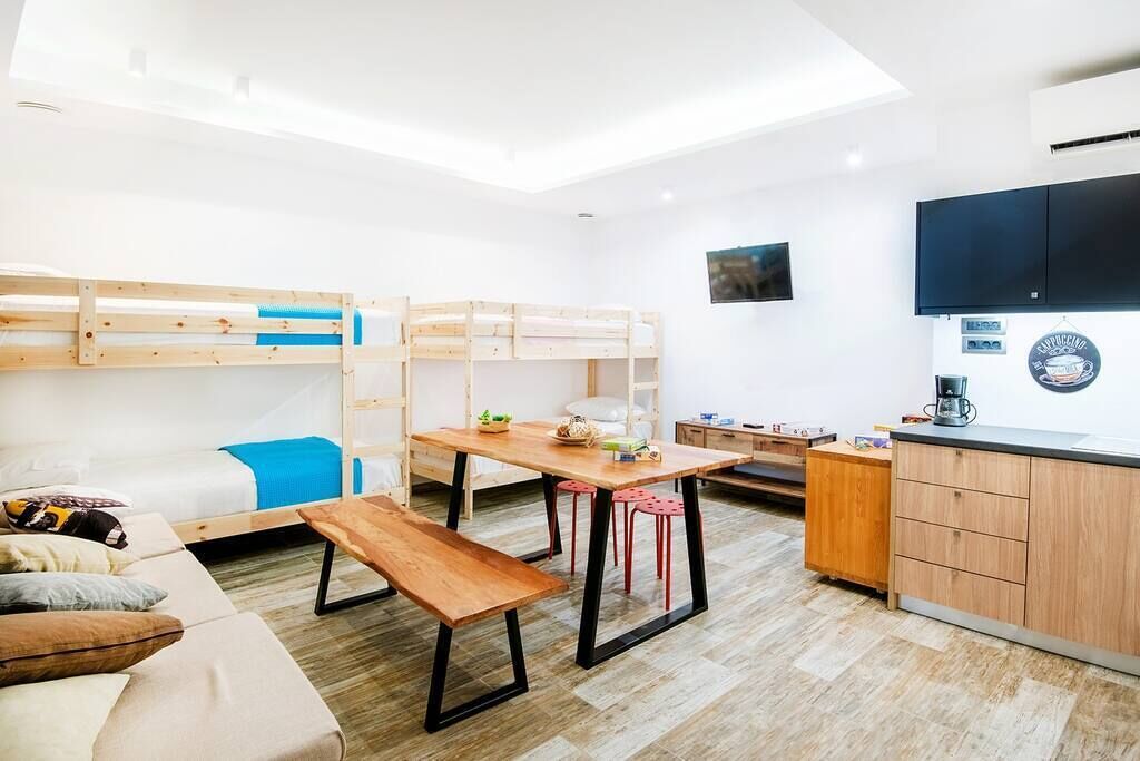 A compact, well-lit room with bunk beds, a wooden table, red stools, a bench, a kitchenette, and a flat-screen TV.