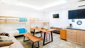 A compact, well-lit room with bunk beds, a wooden table, red stools, a bench, a kitchenette, and a flat-screen TV.