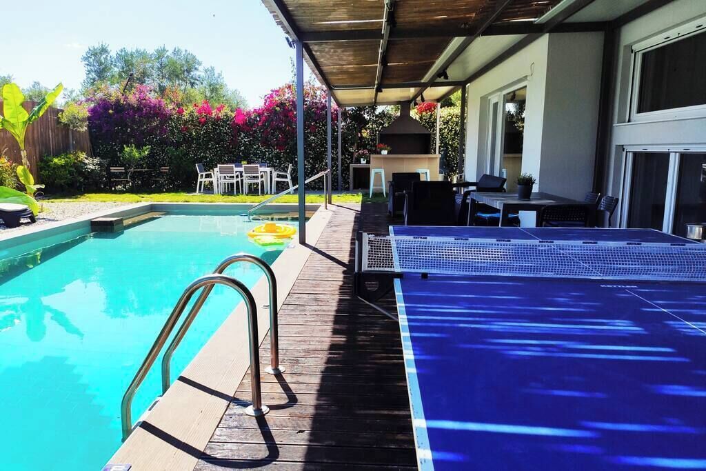 A pool with a wooden deck, a table with chairs, and a barbecue grill.
