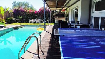A pool with a wooden deck, a table with chairs, and a barbecue grill.