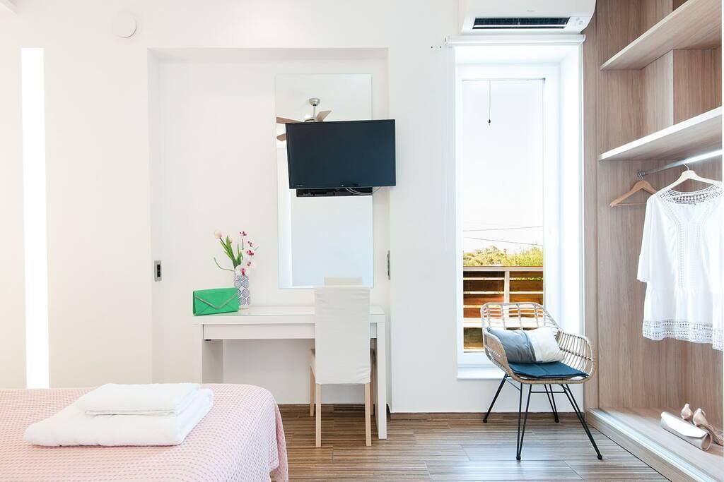 Een hotelkamer met een bed, een bureau met stoel, een aan de muur bevestigde televisie, een raam met uitzicht op groen en een kledingkast met daarin hangende kleding.