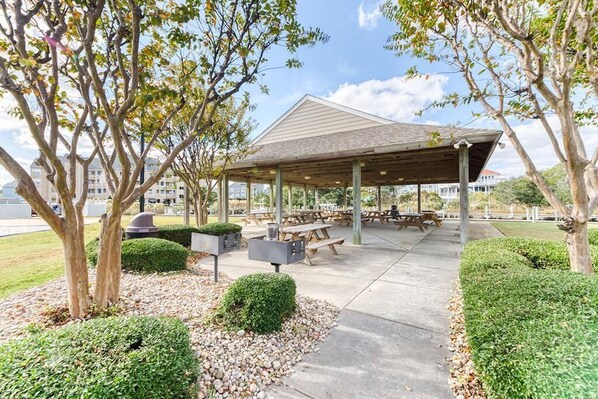 A covered outdoor seating area with picnic tables, surrounded by trees and shrubs.