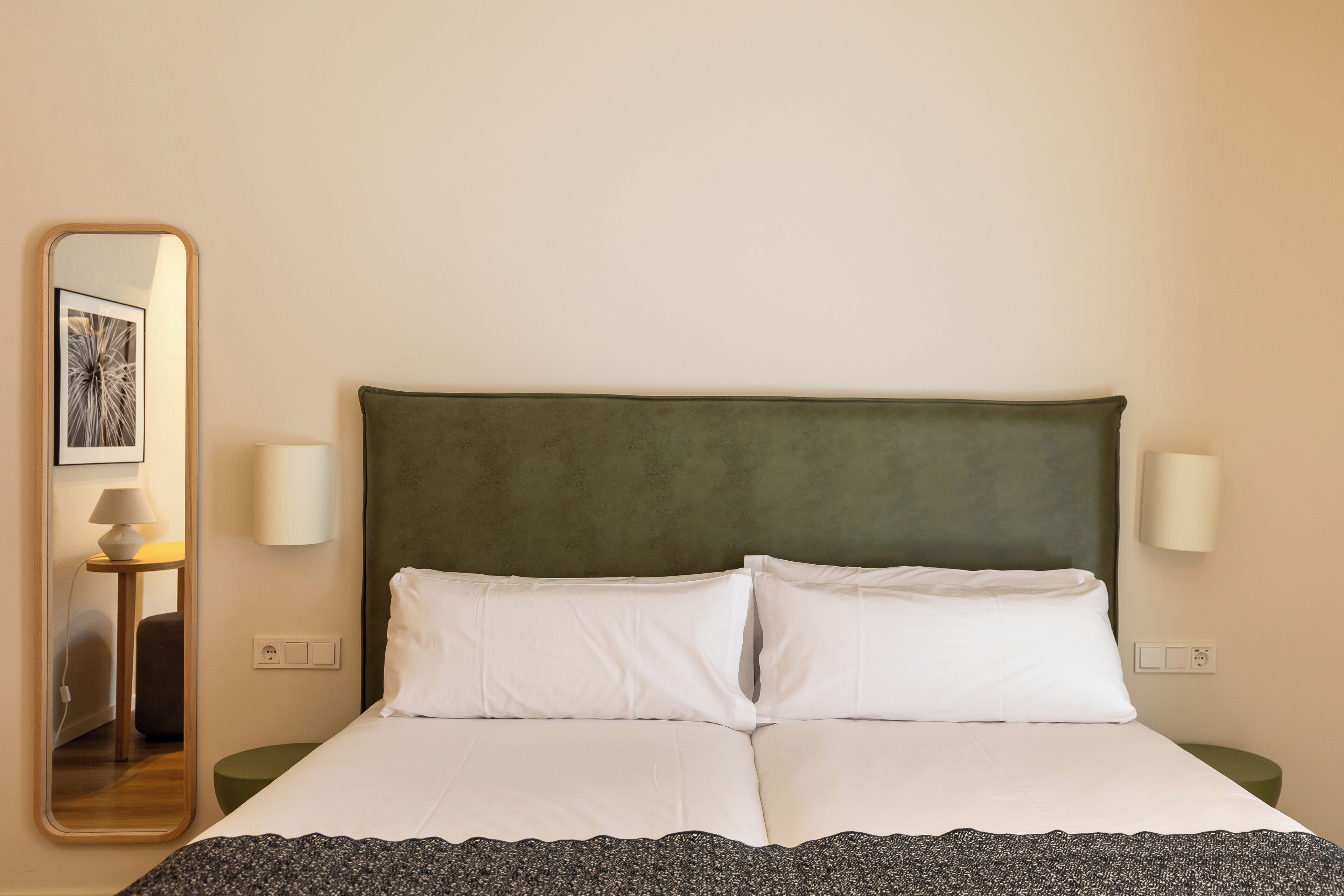 Una cama bien hecha con sábanas blancas y un cabecero verde, dos lámparas de noche y un espejo que refleja parte de la habitación.