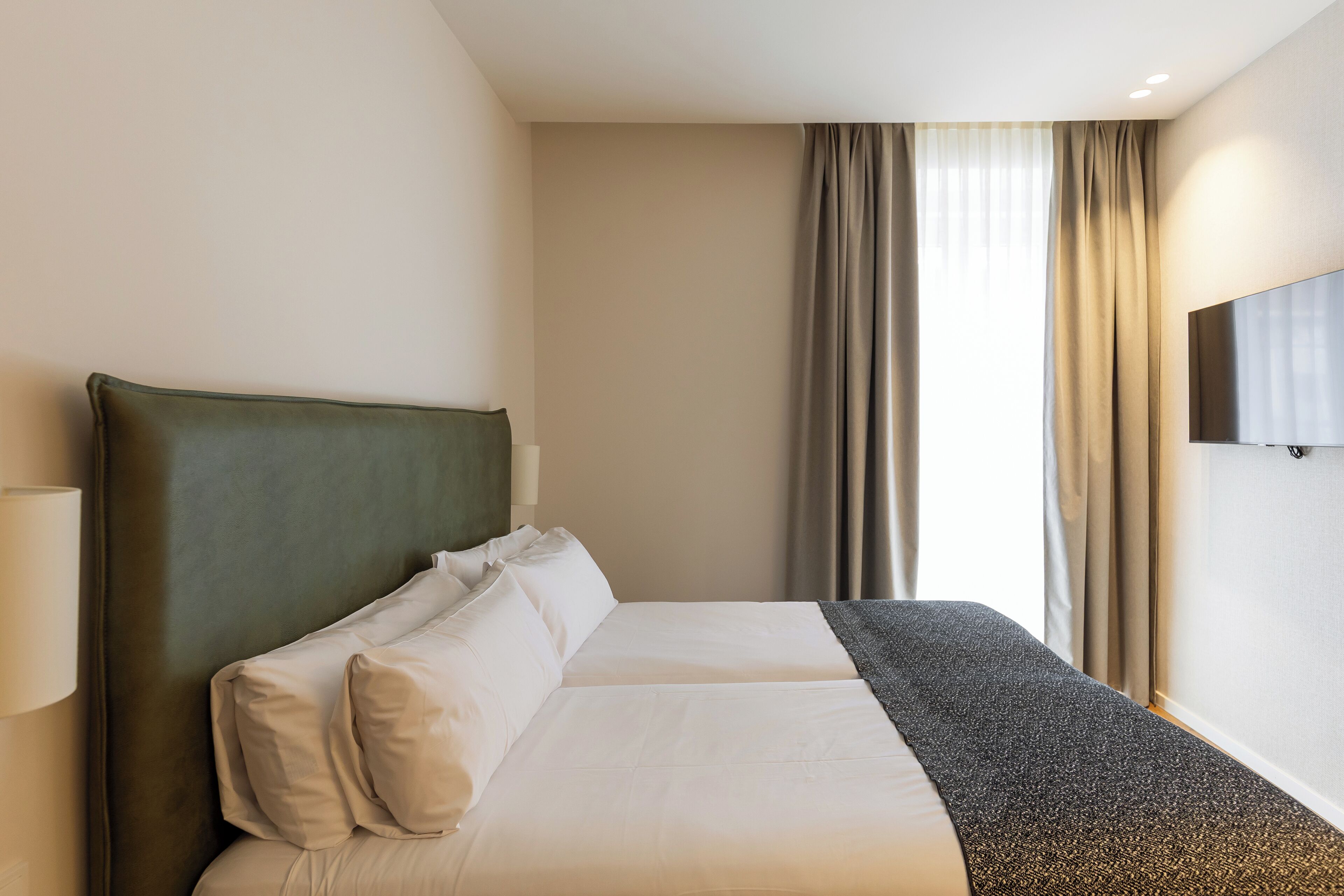 A hotel room with a large bed, a flat-screen TV mounted on the wall, and a window with curtains.