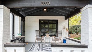 A covered porch with a black ceiling, white brick walls, and a black-framed window. There are two chairs and a small table with a potted plant. - Silver Stallion (Atlanta)