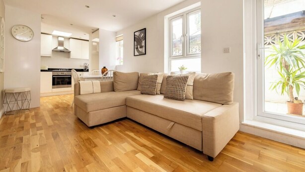 A modern living room with a beige sectional sofa, wooden flooring, a kitchen in the background, and a large window with a view of greenery. - The Queenstown Villa (London)