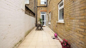 A narrow alleyway with a tiled patio, a table with chairs, and a potted plant. - The Queenstown Villa (London)