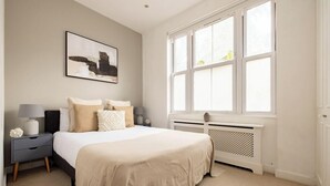 A bedroom with a large bed, a nightstand, a lamp, a radiator, and a window. - The Queenstown Villa (London)