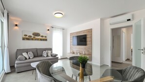 Een moderne woonkamer met een glazen eettafel, een grijze bank en een flatscreen televisie die aan een houten wand is bevestigd.