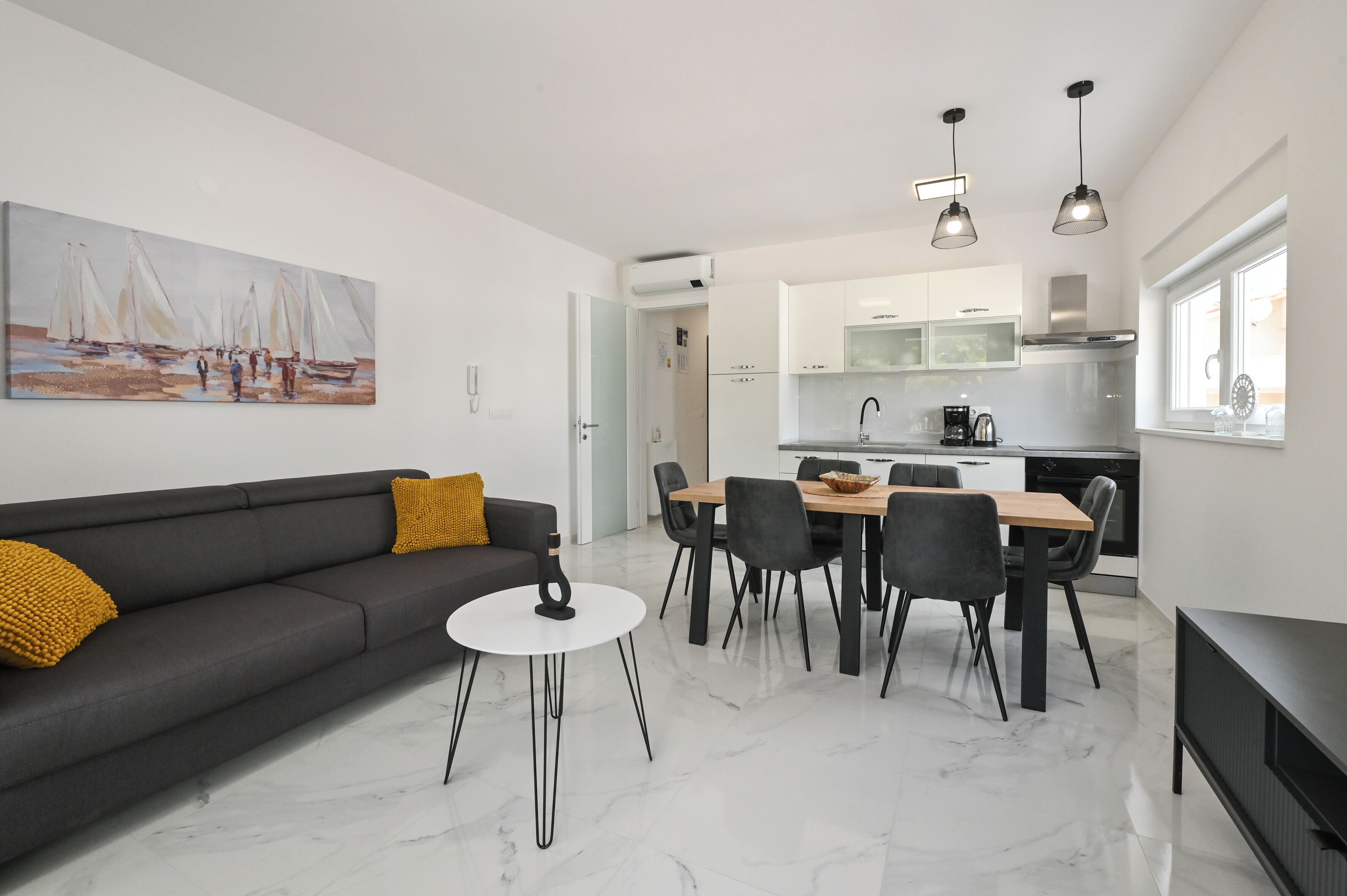A modern living room with a sofa, coffee table, and a dining area with chairs and a table. There is a kitchen in the background with cabinets and a window.